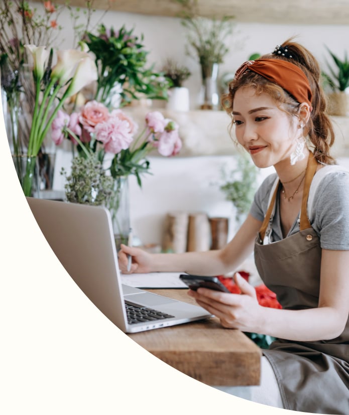 Asian female florist using smartphone while working on laptop